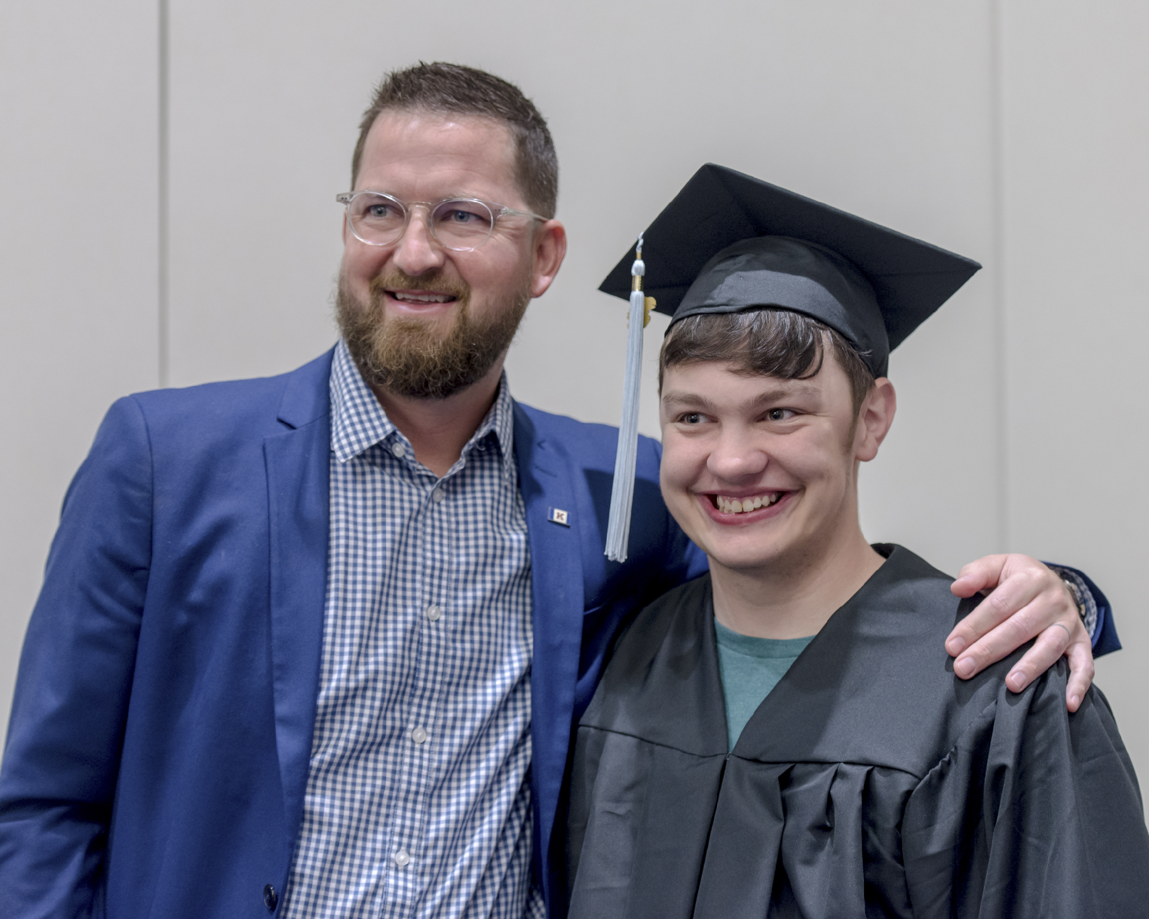 "An older man with a beard and glasses stands with his arm around a younger man wearing graduation cap and gown while both smile"