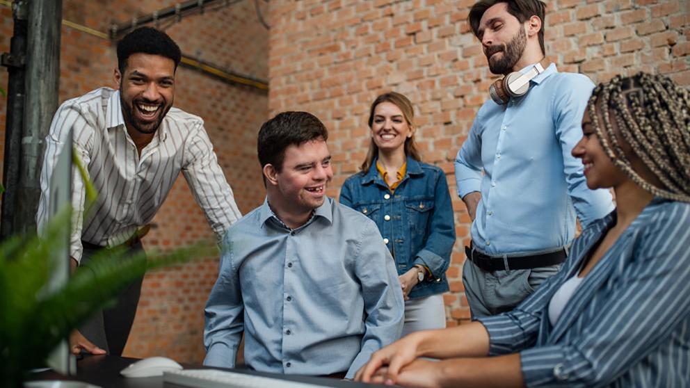 A man wearing a blue button up shirt who appears to have Down syndrome sits in an office environment with coworkers surrounding him as they smile together