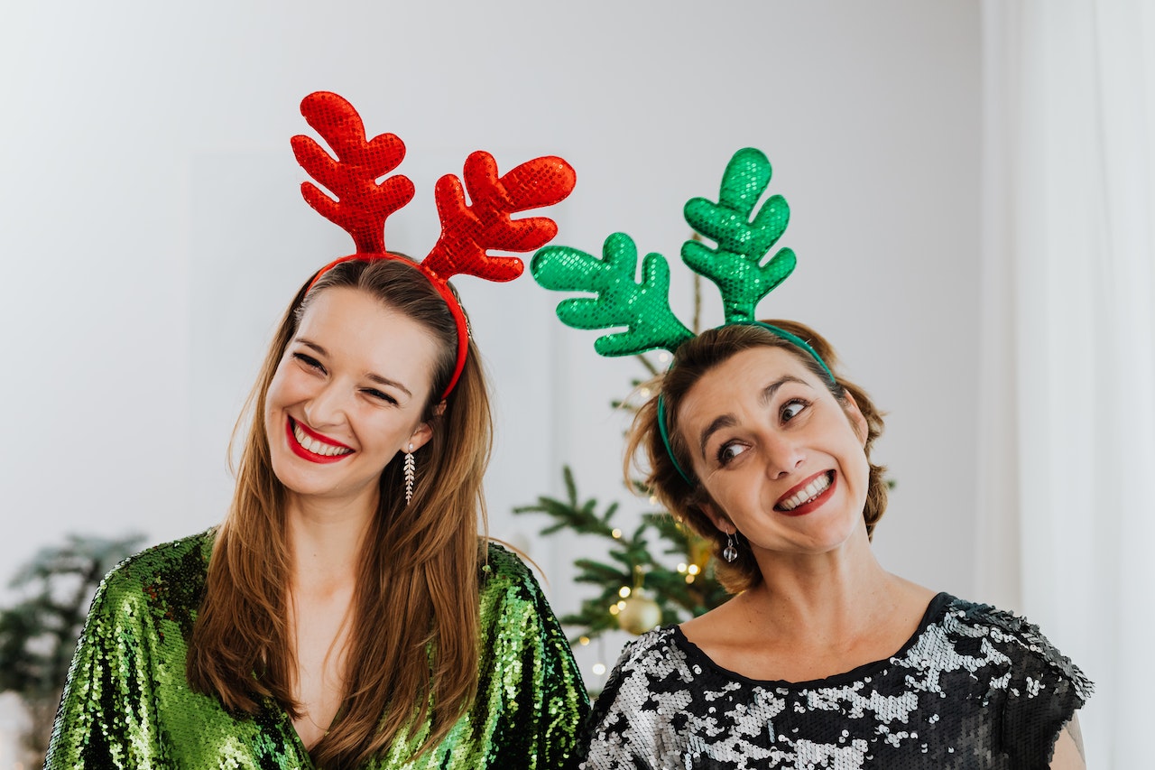 Two women with sequin dress and costume antlers on their heads smile with a Christmas tree in the background