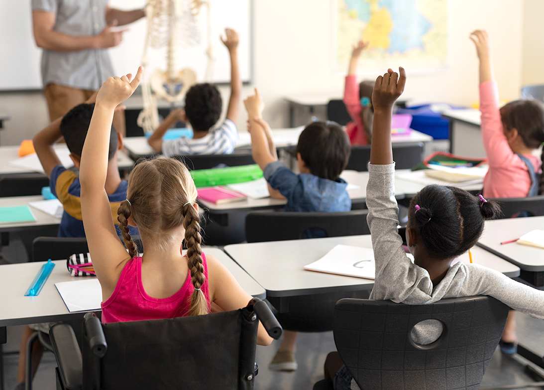 "A diverse classroom of students, including a girl in a wheelchair, face the front of a classroom with hands raised, looking toward their teach who is standing next to a human skeleton model"