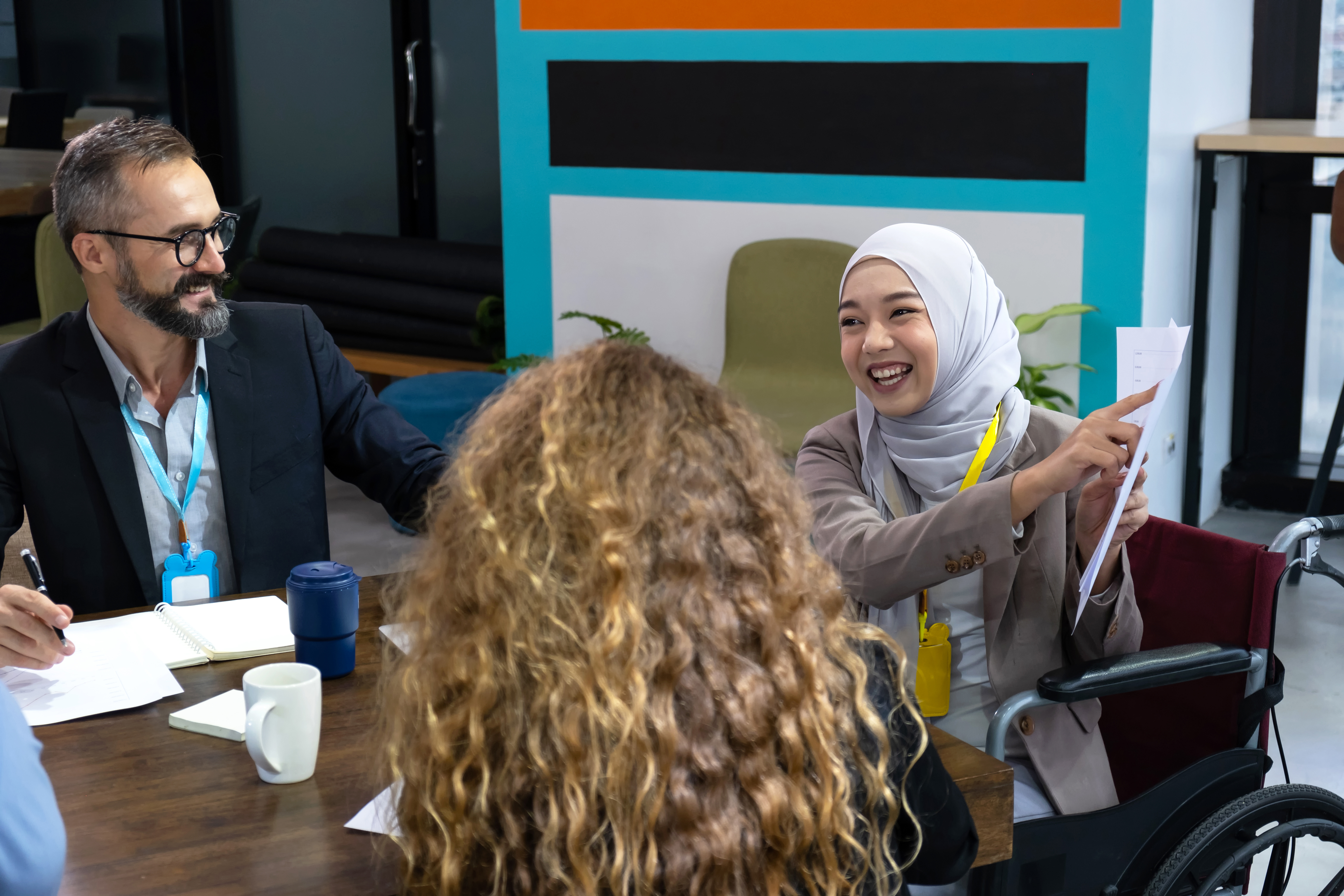 "A woman with a white hijab covering her hair smiles while pointing to a piece of paper while seated in a wheelchair at a conference table with other individuals seated nearby look at her report"
