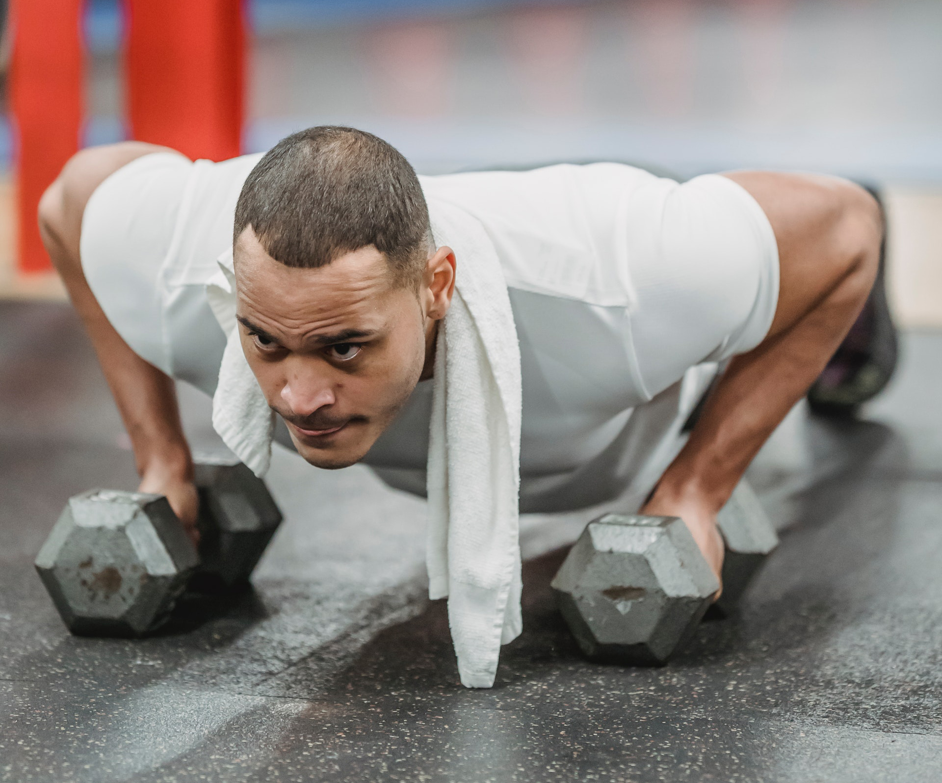 "A man wearing a white shirt with short hair holds weights in his hands while doing pushups on the floor of a gym."