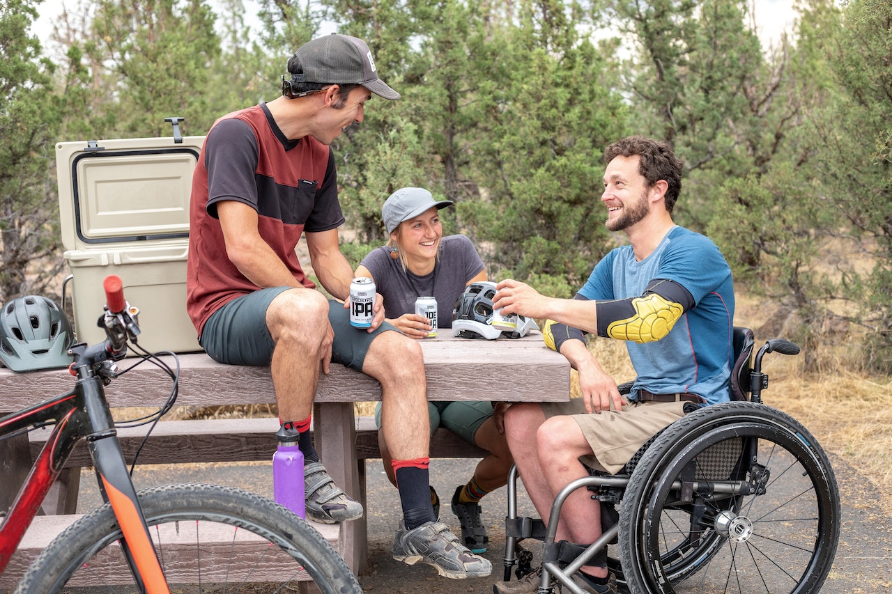 "Three young adults, including one with a wheelchair and another next to a bike, sit outdoors at a picnic table in the outdoors"