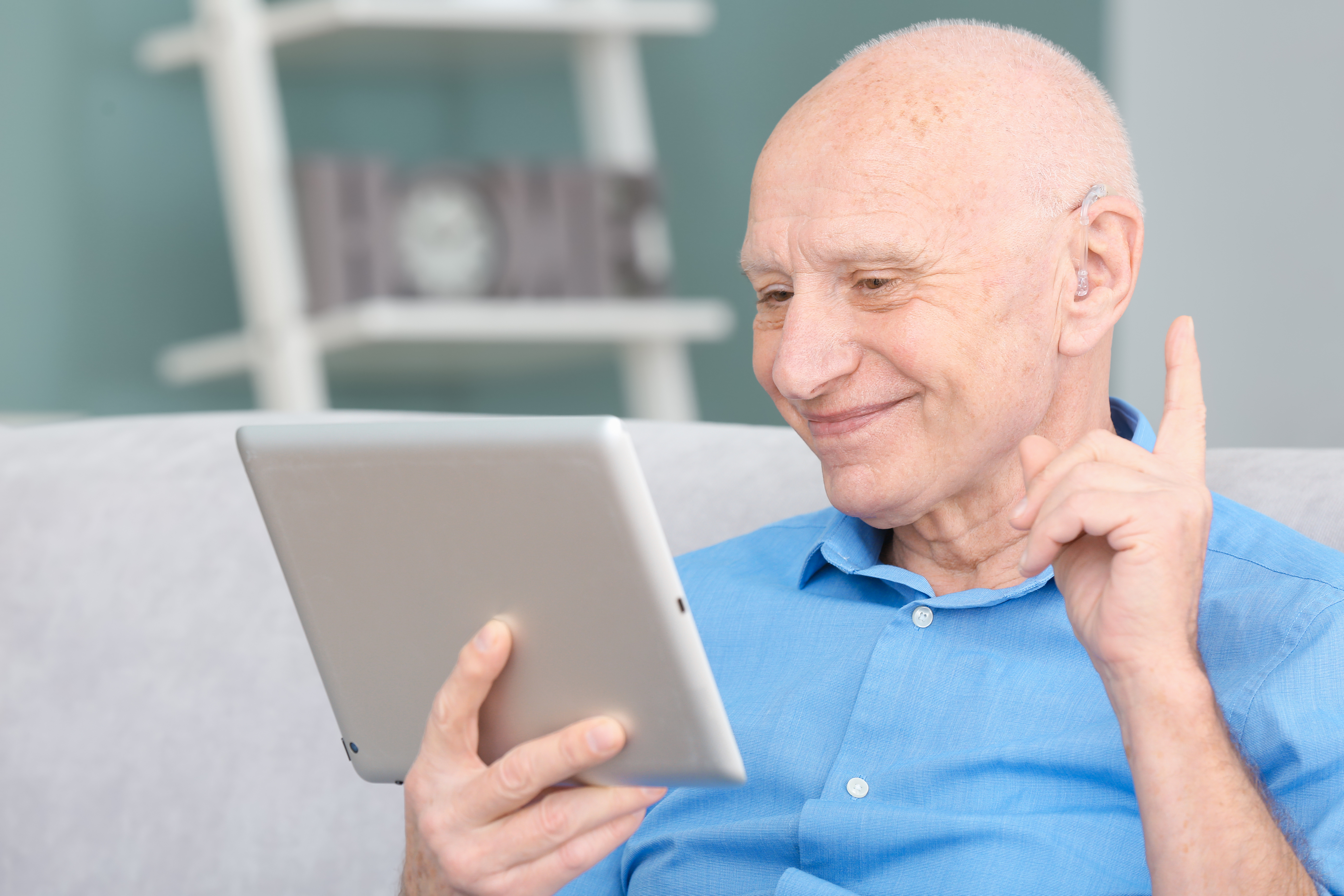 "An older man in a blue shirt with a hearing aid smiles as he looks into an iPad screen and holds up his index finger"