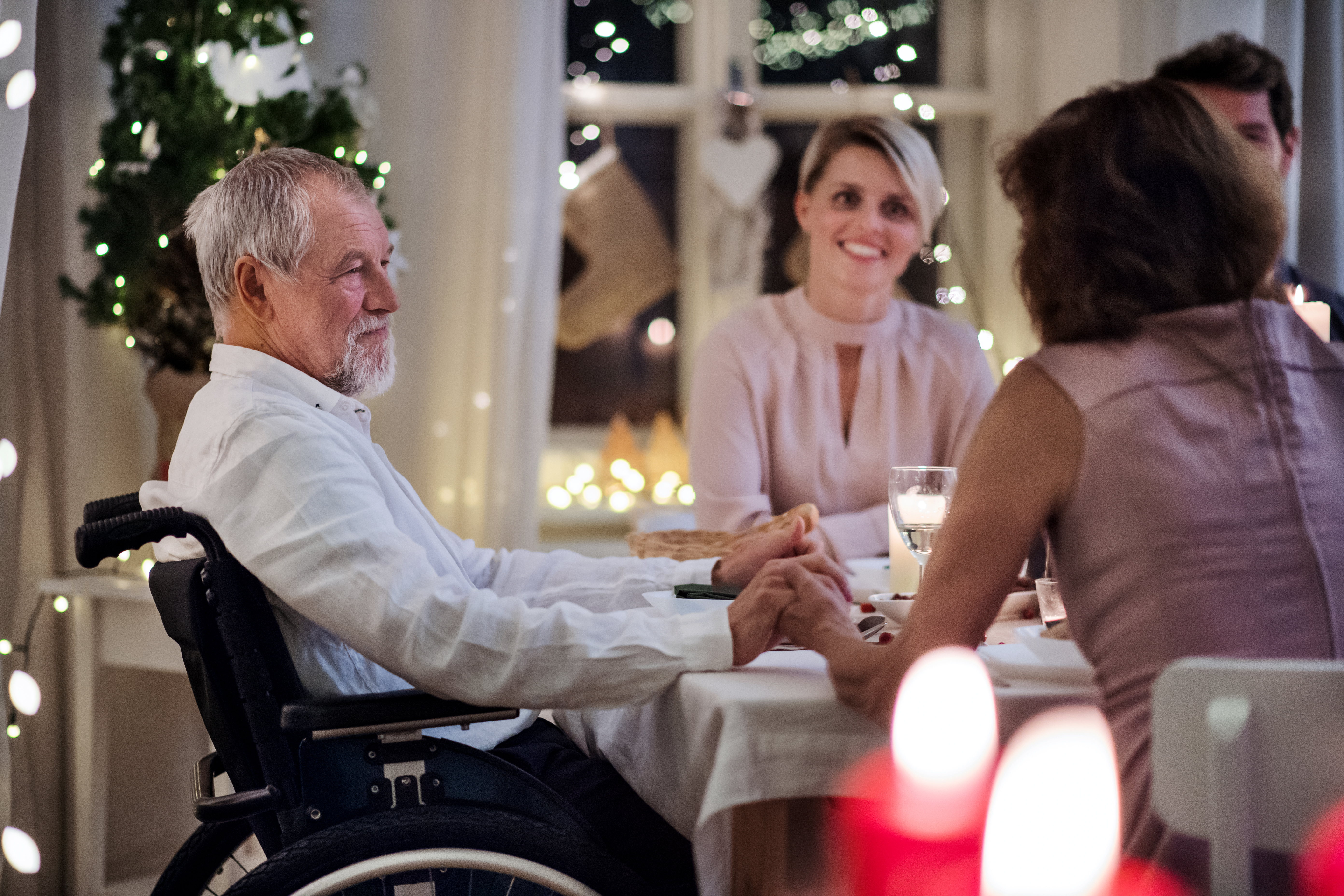 "A man with white hair and a beard sits in a wheelchair at a dinner table and holds with a woman sitting next to him who is speaking to other people sitting with them in a home decorated for Christmas"