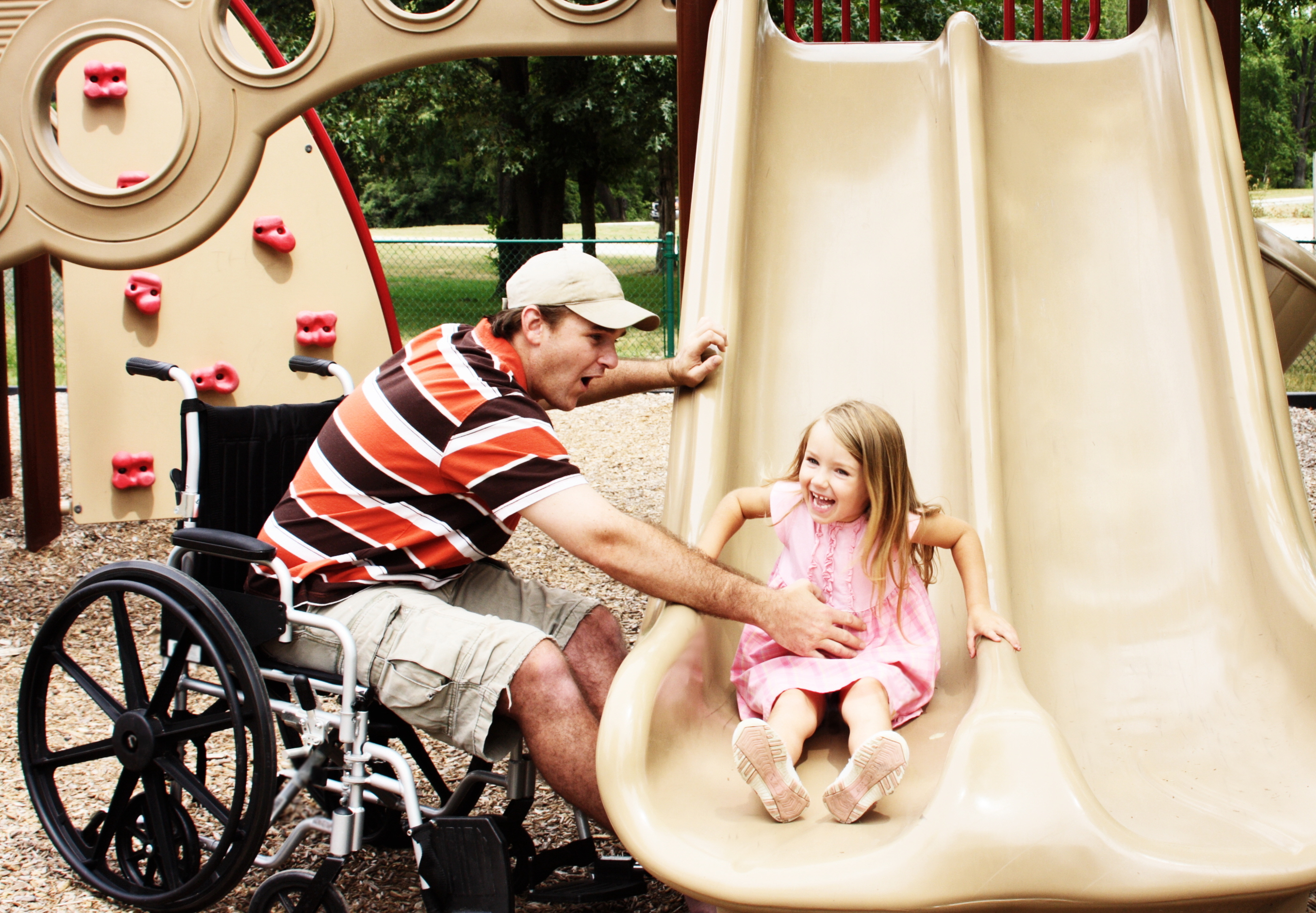 "A man sitting in a wheelchair leans toward a slide at a playground to help catch a young girl smiling as she slides down"