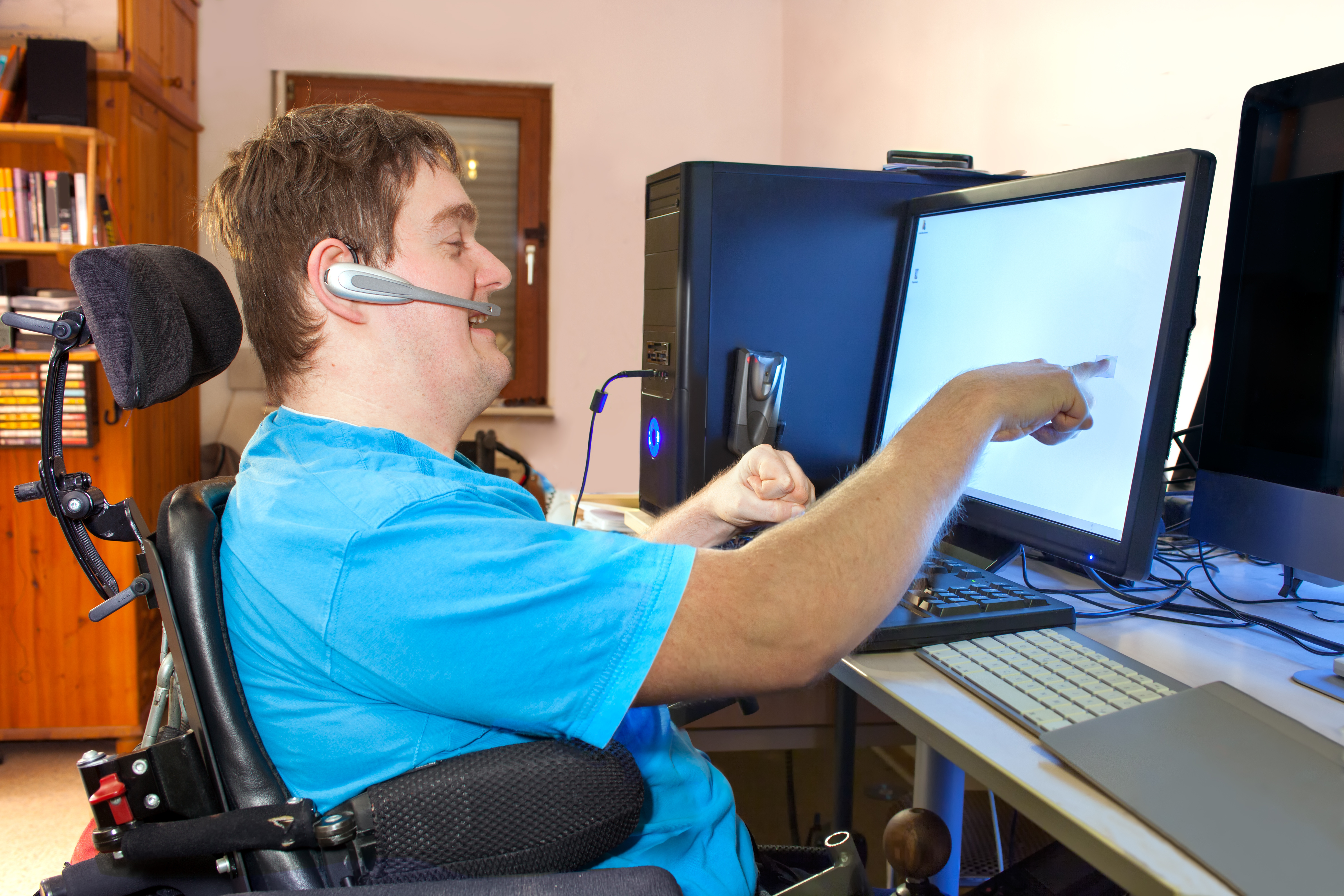 "A young man wearing a blue t-shirt sitting in a wheelchair uses a computer"