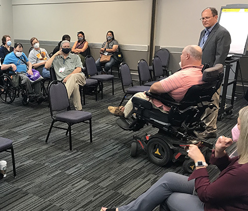 A seated audience is facing researcher Brad Linnenkamp, seated in power chair, and researcher Evan Dean, standing as they present information at the Kansas Disability Caucus. 