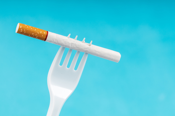 A plastic fork is holding a cigarette; the background is a solid light blue