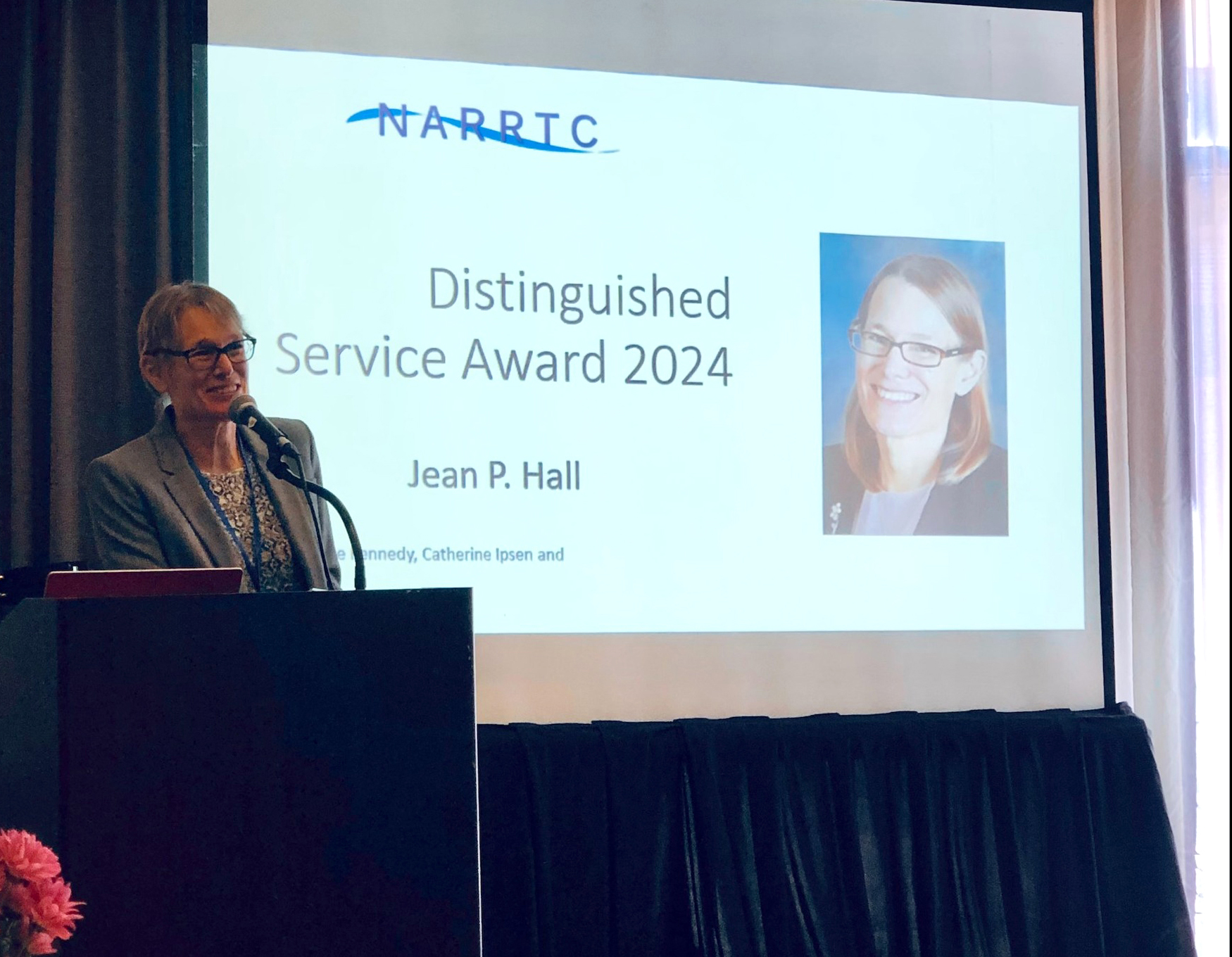 "Jean Hall stands at a podium in front of a screen with her name and image on it"