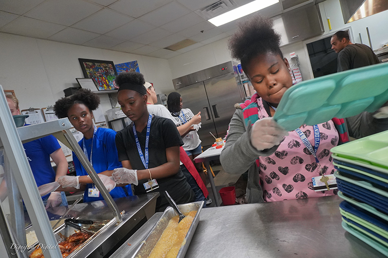 A group of teens participating in an out of school program. They are in a cafeteria kitchen stacking lunch trays.