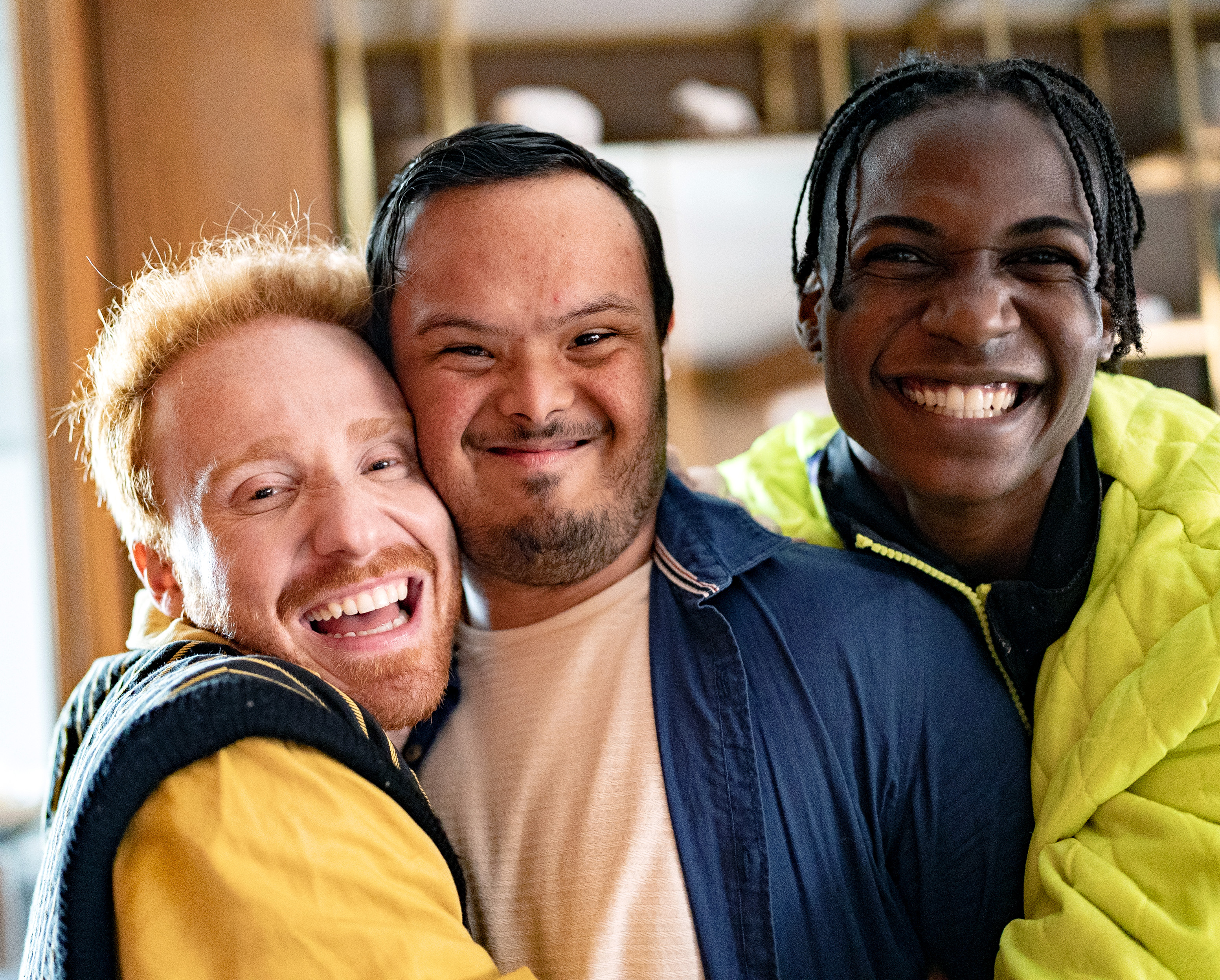 "A man with red hair and a beard and a man with dark hair and a beard who has Down syndrome and a man with black hair and braids smile while in an embrace as they pose for a photograph"