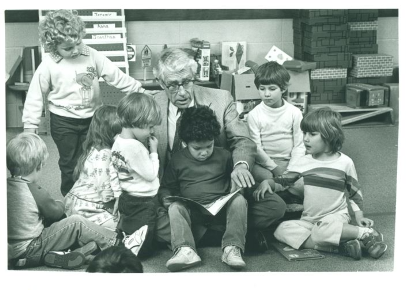 "Richard L. Schiefelbusch reads to a small group of children while seated on a floor in a playroom"