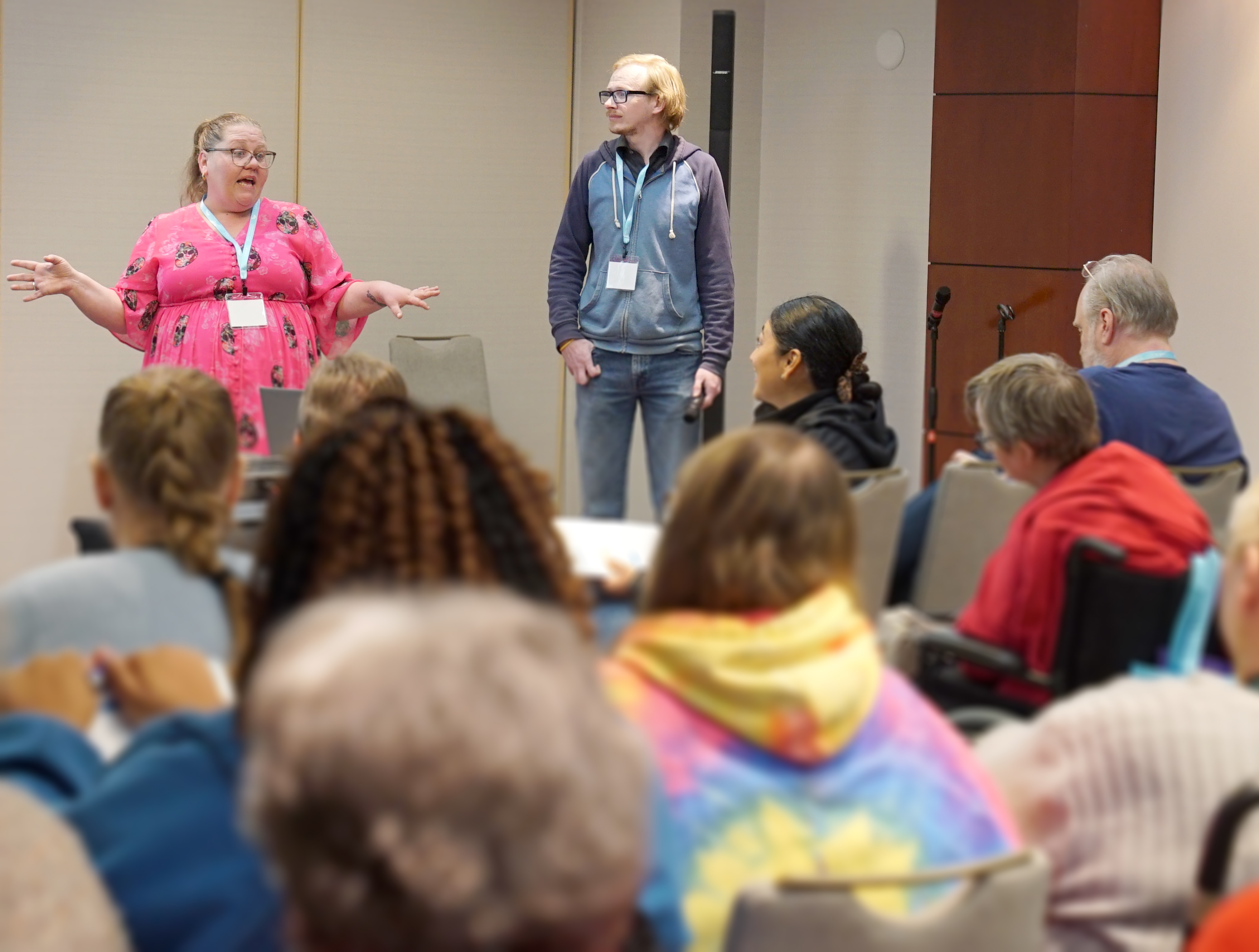 "A woman with blonde hair in a ponytail wearing glasses and a pink dress stands next to a man wearing glasses and a blue sweatshirt while speaking to a crowded audience"