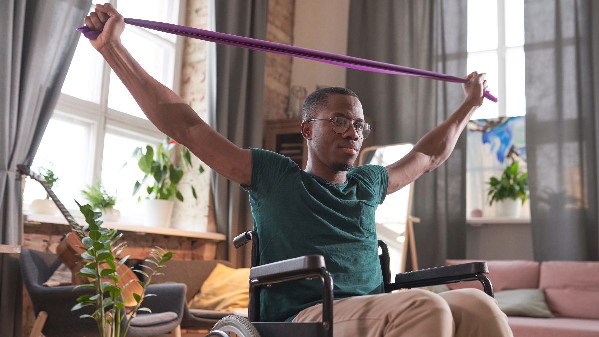 "A young man with short black hair and glasses uses a resistance band to exercise his arms while sitting in a wheelchair in his home"