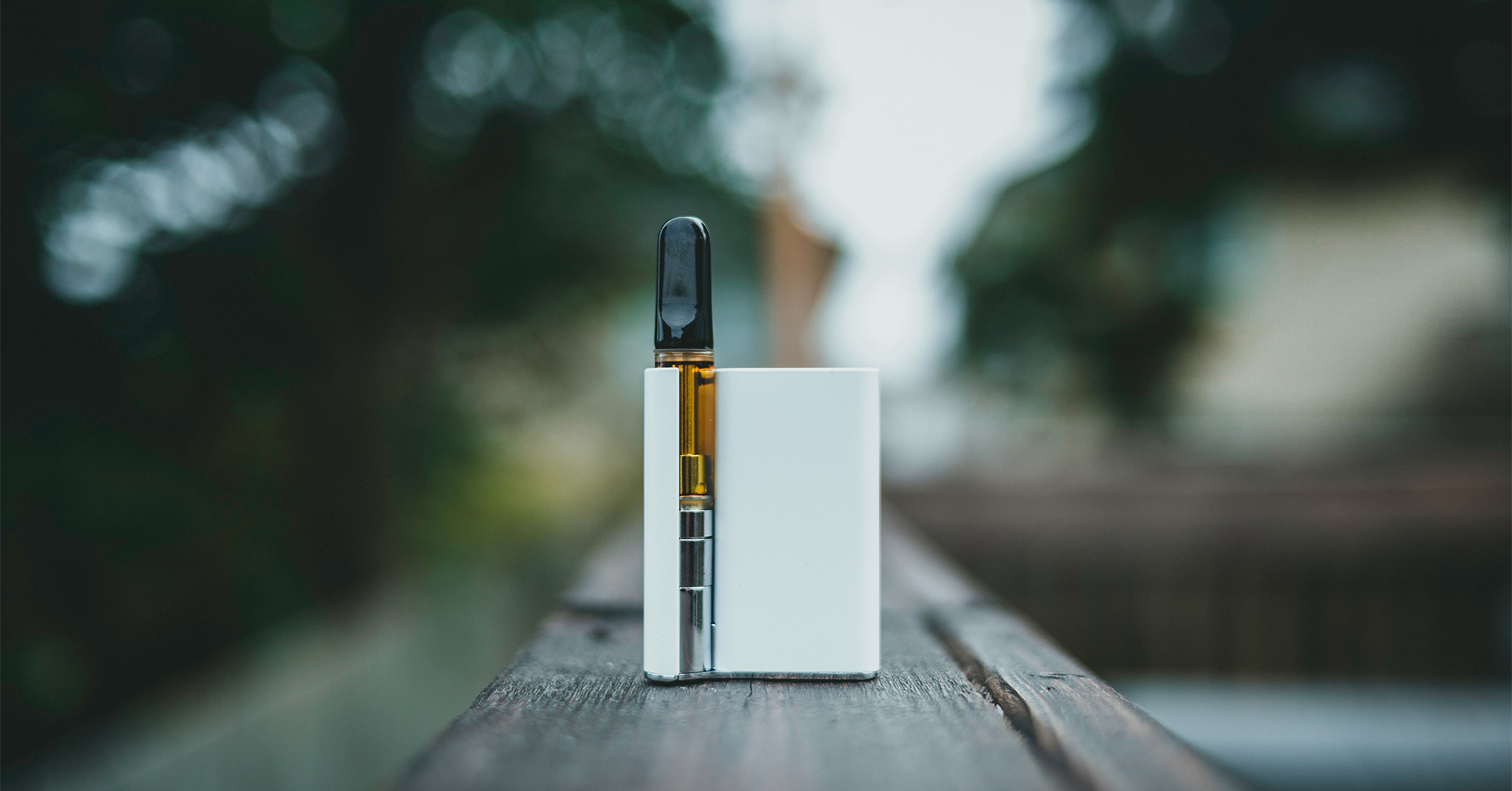 A vaping device is in focus in the center of a wood table outside with filtered sunlight through trees.