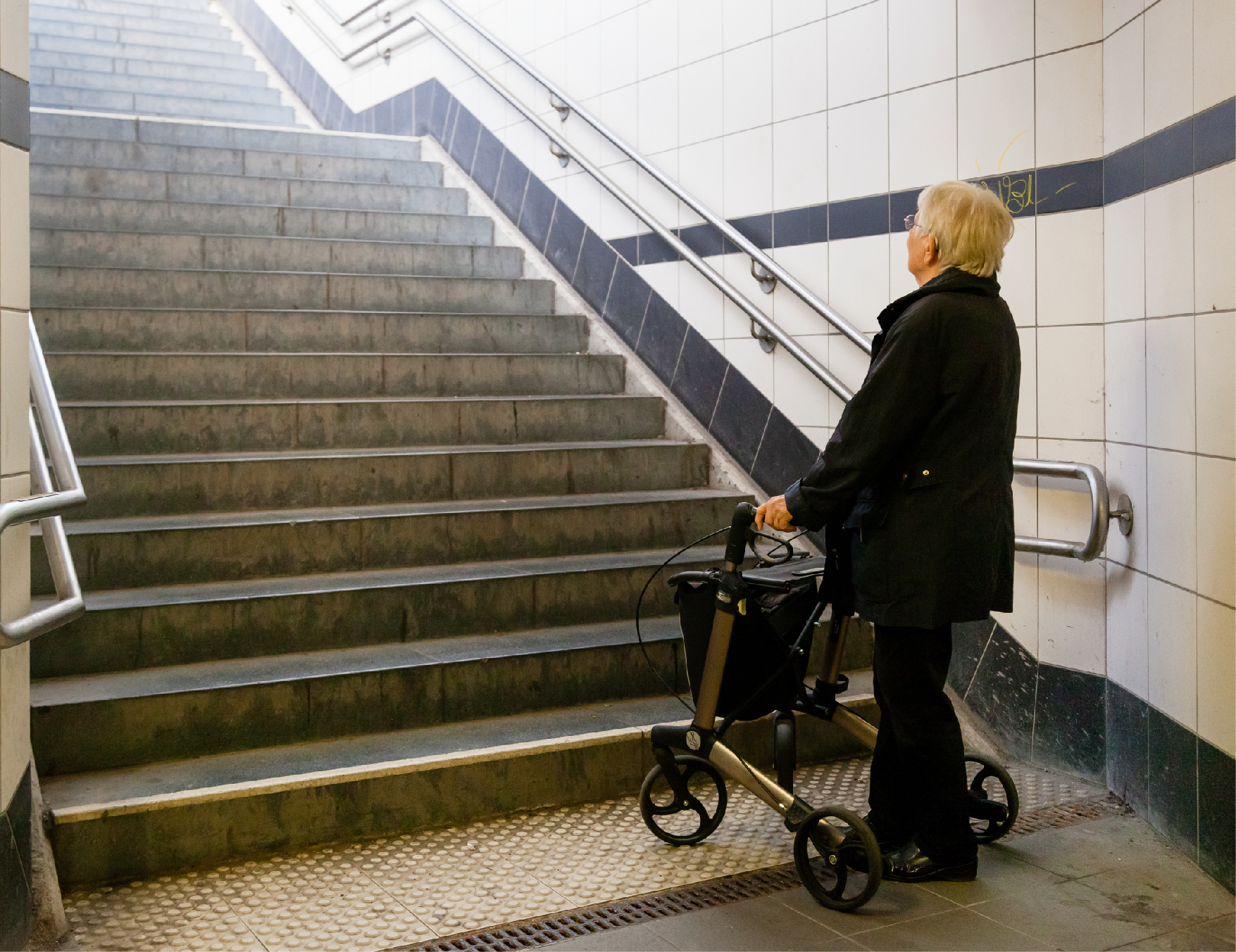 "A woman with short white hair stands with help from a walker looking up at a steep flight of stairs"