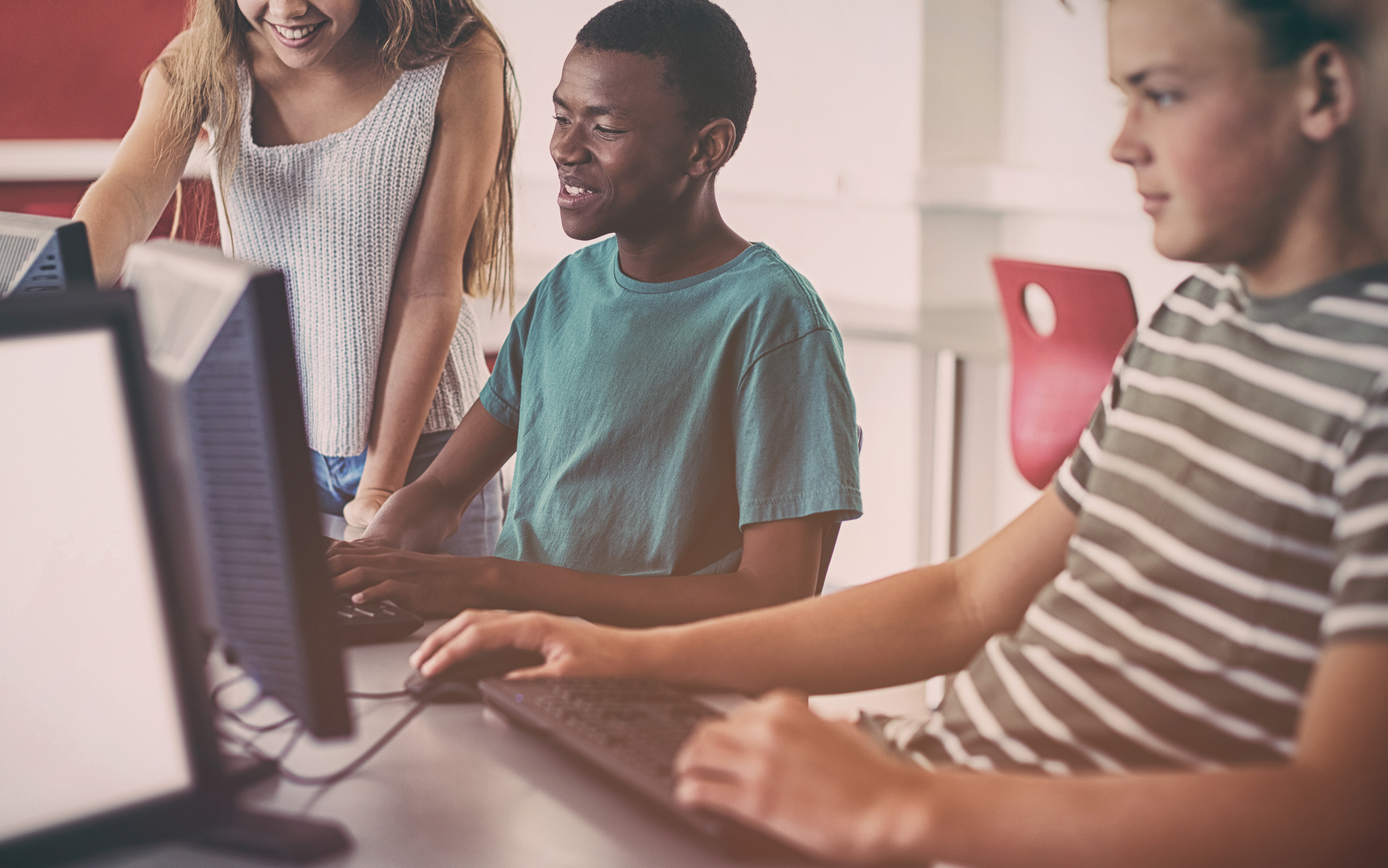 "Two teen boys sit and a teen girl stands while they work at computers in a classroom"