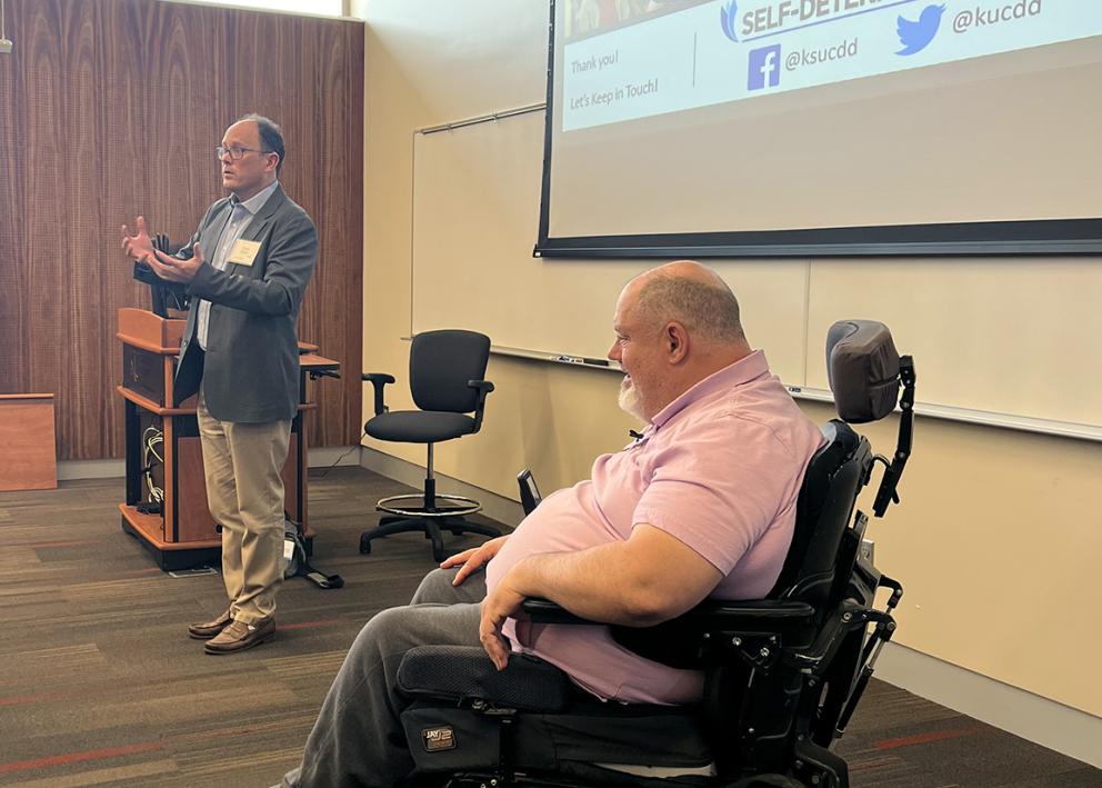 A man standing at the head of a classroom speaks into a microphone as a second speaker seated in a wheelchair reacts
