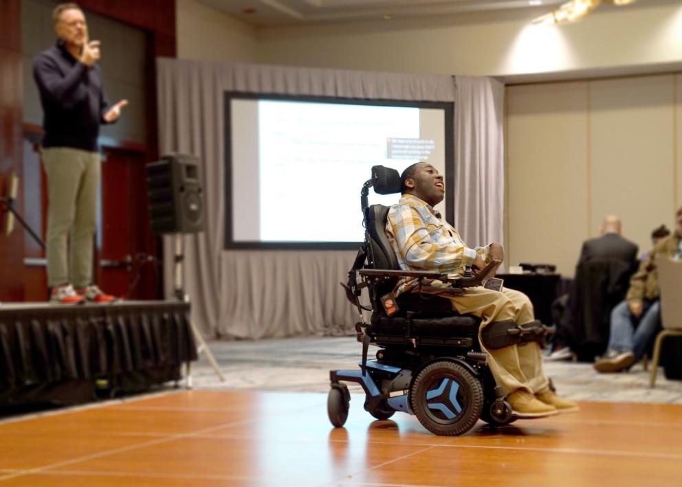 A man with short black hair speaks to an audience in a large room while seated in a motorized wheelchair in front of a stage