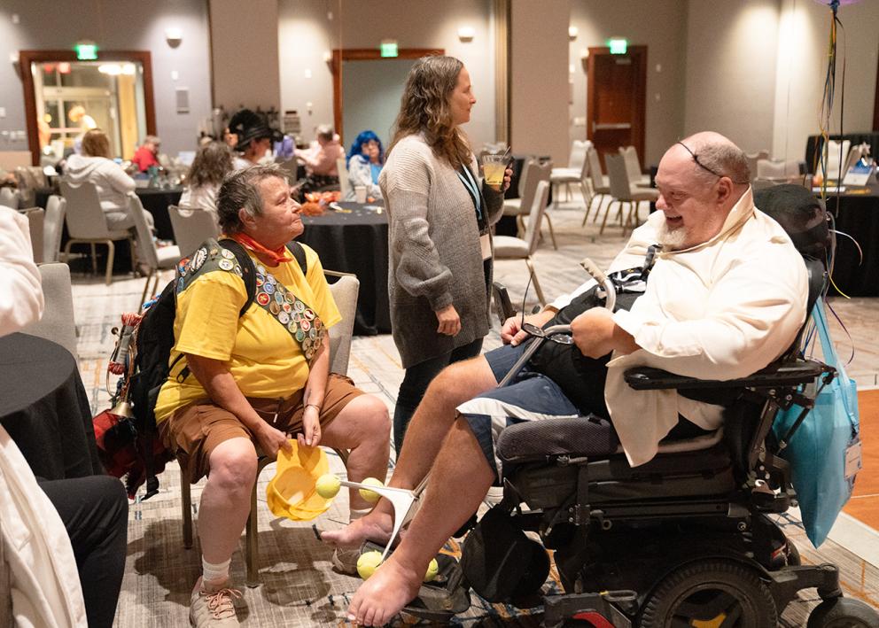 A person seated in a yellow shirt with a Scout sash decorated with badges sits at a chair next to a man wearing a white cape while seated in a motorized wheelchair
