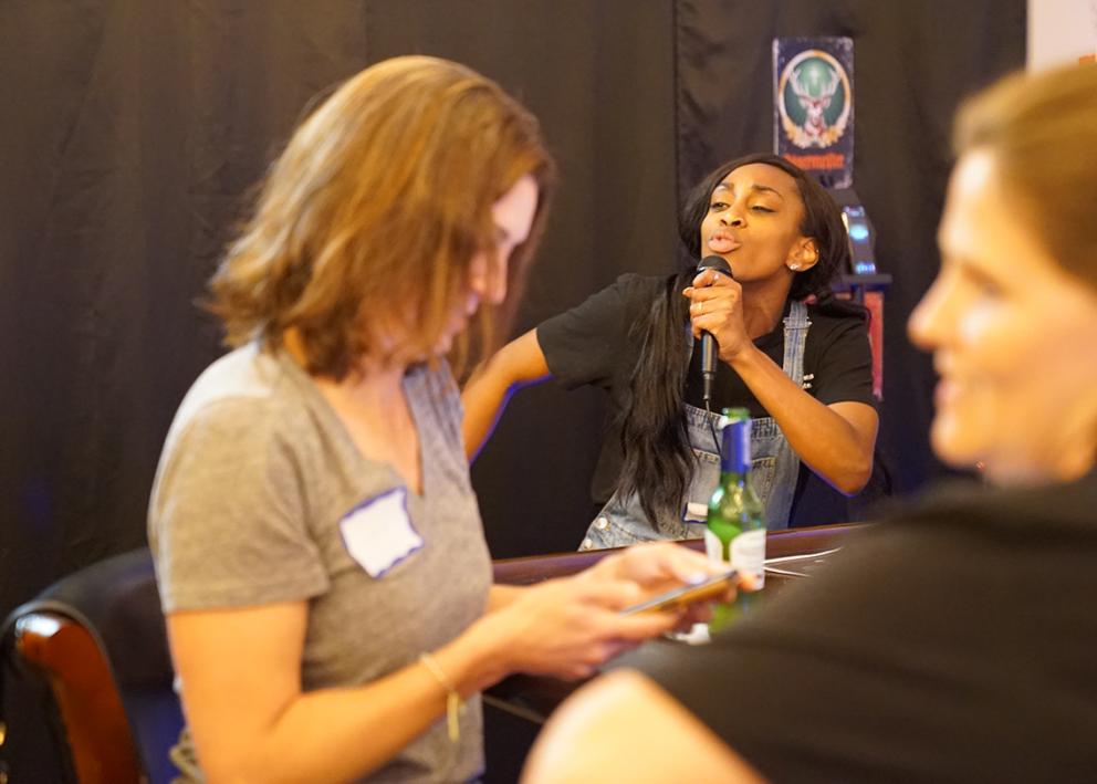 A woman sings into a microphone in the background while a woman checks her phone nearby