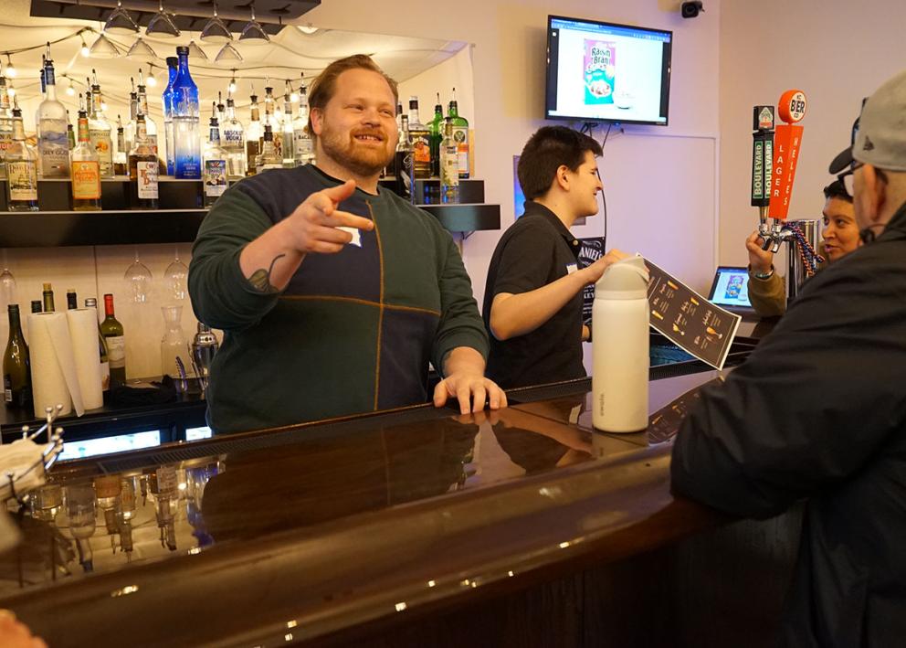 A man working as a bartender gestures to people sitting at the bar 