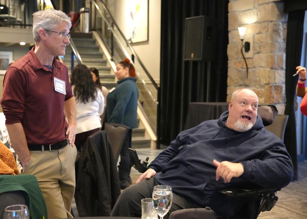 A man with gray hair and a burgundy collared shirt leans back while standing to the left of a man with a white beard and a blue sweatshirt seated in a wheelchair who is speaking and lookup up to the right