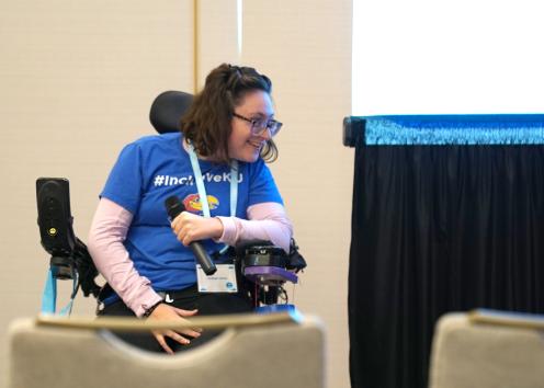 A woman wearing a blue shirt with a KU Jayhawk logo holds a microphone while seated at the front of a room in her motorized wheelchair