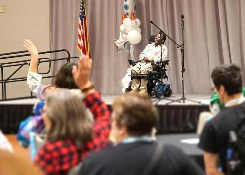 A man seated in a motorized wheelchair on stage faces audience members seated at tables raise their hands
