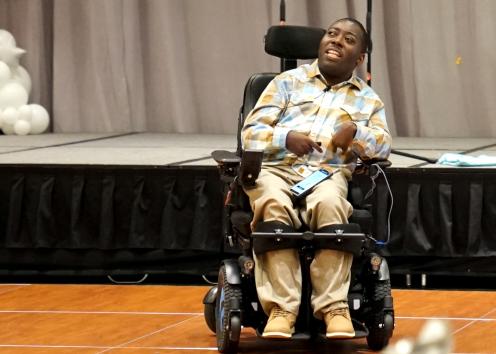 A man with short black hair speaks while seated in a motorized wheelchair in front of a stage