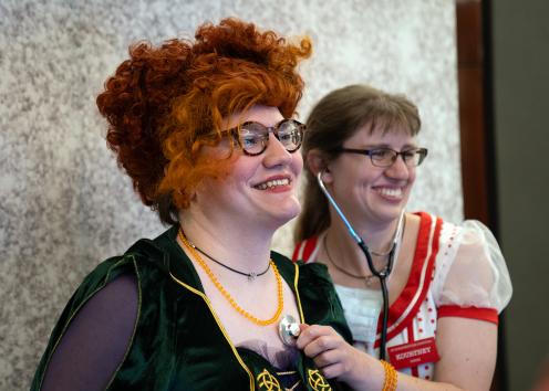 A woman with red hair and glasses wearing a black dress stands with a woman holding a stethoscope to her chest who has on a red and white costumed dress