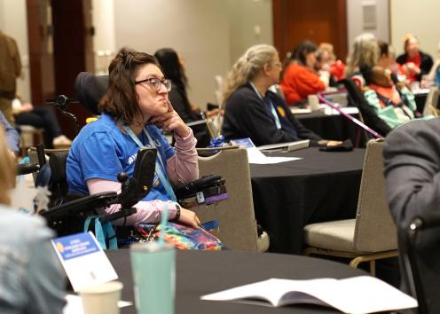 A woman with brown wavy hair wearing glasses and a blue shirt places a finger on her chin while listening from her seat in her motorized wheelchair