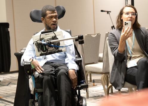 A man with short black hair and a beard sits with a screen device that is attached to his wheelchair next to a woman with brown hair and glasses who is speaking while holding a phone