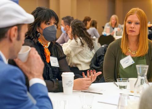 People at a table listen as a woman wearing a black face mask gestures while speaking