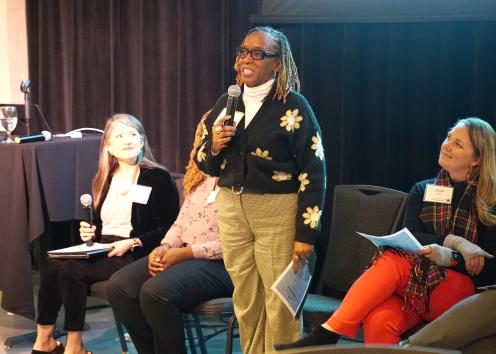 A woman with black glasses and braided hair wearing a black knit sweater with yellow flowers smiles while speaking into a microphone while a group of women seated next to her listen to her speak
