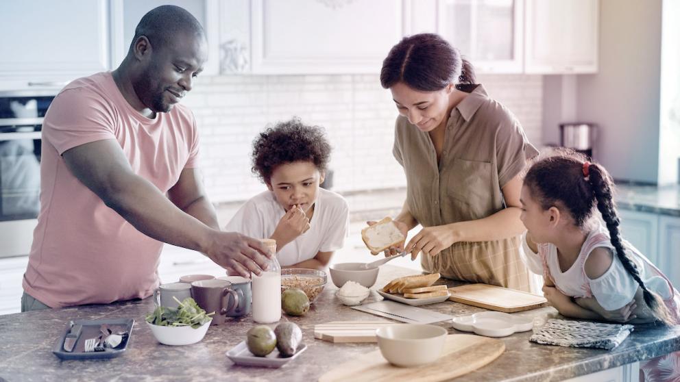 Stock image of man, woman, two children gathered around kitchen island with wooden countertop, preparing meal with sliced bread.