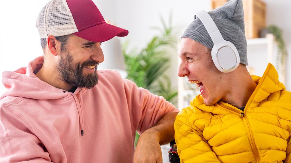 A man wearing a ball cap and a pink hoodie smiles as he communicates with his friend who has ALS and is wearing a yellow jacket and headphones
