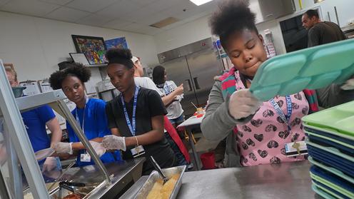 A group of teens participating in an out of school program. They are in a cafeteria kitchen stacking lunch trays.